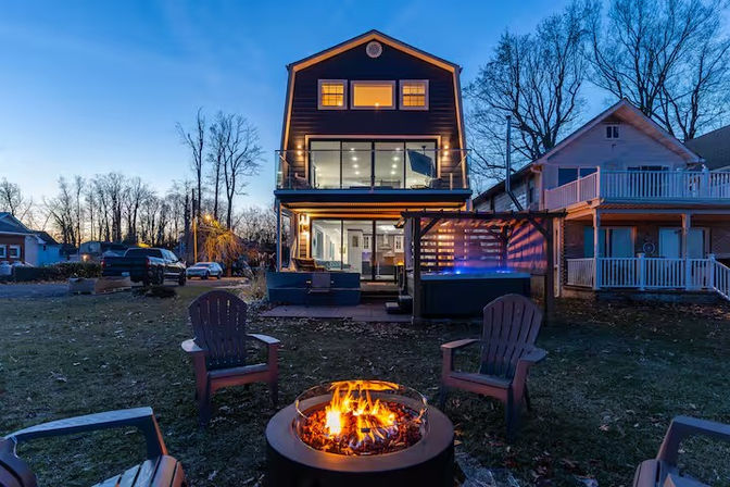 Cozy three-story modern home at dusk with illuminated glass balcony, backyard fire pit surrounded by Adirondack chairs, patio seating and hot tub.