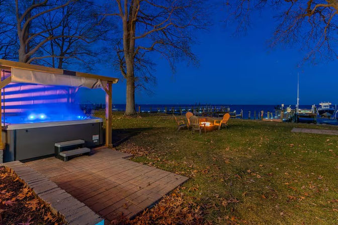 Evening waterfront backyard by a lake: blue-lit hot tub under a pergola, cozy fire pit with chairs, dock and moored boats