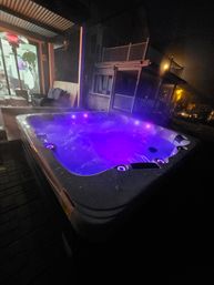 Purple-lit outdoor hot tub bubbling on a covered backyard patio at night, steam rising with neighboring two-story homes and a lit interior room visible