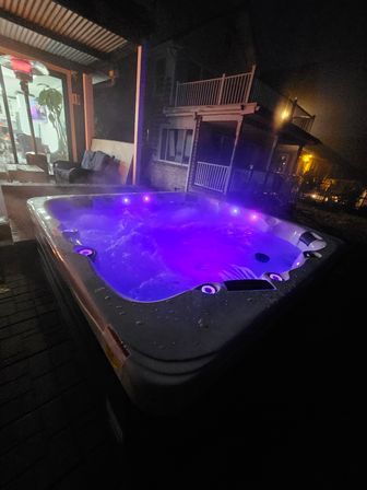 Purple-lit outdoor hot tub bubbling on a covered backyard patio at night, steam rising with neighboring two-story homes and a lit interior room visible
