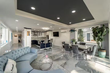Sunlit open-concept modern coastal living area with a tufted light-blue curved sofa, glass coffee tables, glossy marble-look floors, navy recessed ceiling, dining table with grey upholstered chairs, kitchen island with bar stools, wall-mounted TV and large potted plant.