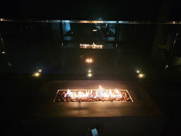 Modern outdoor patio gas fire pit with glowing lava rocks and dancing flames, reflected in a glass railing on a dark night