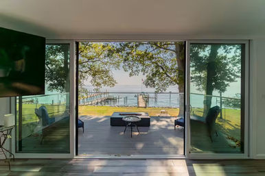 Sunlit lakefront living room with open sliding glass doors to a deck featuring chairs and a fire pit table, grassy yard and wooden dock extending into a calm lake framed by leafy trees.