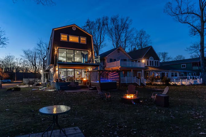 Dusk view of a three-story modern house with glowing balconies and large windows, neighboring homes, and a cozy backyard fire pit with Adirondack chairs on a grassy yard beneath bare trees.