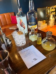 Craft cocktail station on a wooden bar: two spirit bottles, small vials of citrus and honey, a rocks glass of crushed ice topped with a dried citrus wheel, and a printed 'Eat a Peach' recipe card.