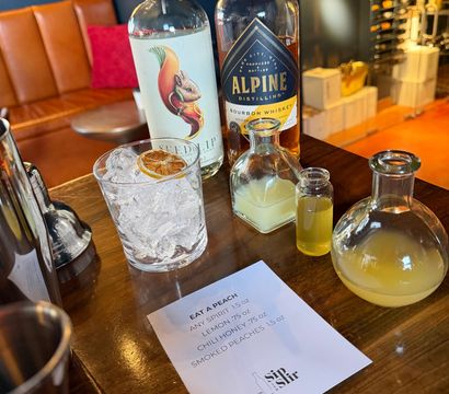Craft cocktail station on a wooden bar: two spirit bottles, small vials of citrus and honey, a rocks glass of crushed ice topped with a dried citrus wheel, and a printed 'Eat a Peach' recipe card.