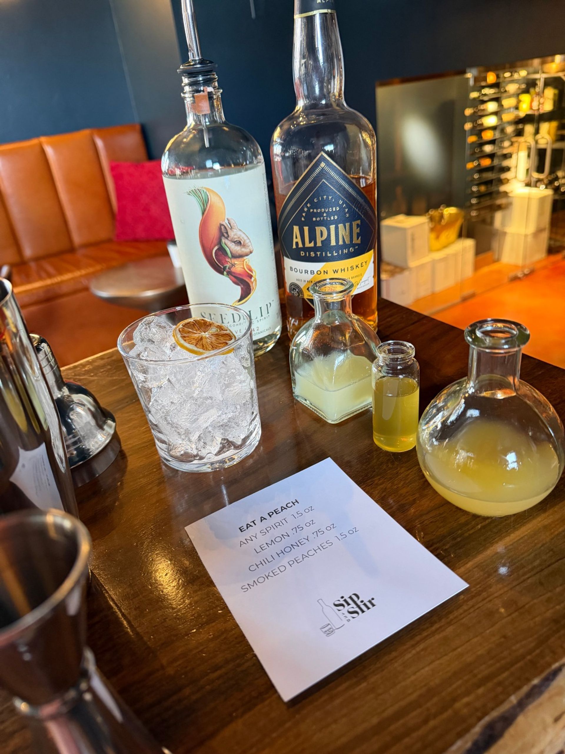 Craft cocktail station on a wooden bar: two spirit bottles, small vials of citrus and honey, a rocks glass of crushed ice topped with a dried citrus wheel, and a printed 'Eat a Peach' recipe card.