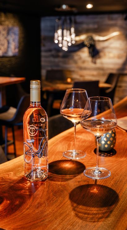 Bottle of rosé with a bicycle label and two poured wine glasses on a polished wooden bar in a cozy, dimly lit wine bar with wood-paneled wall and decorative longhorn in the background.