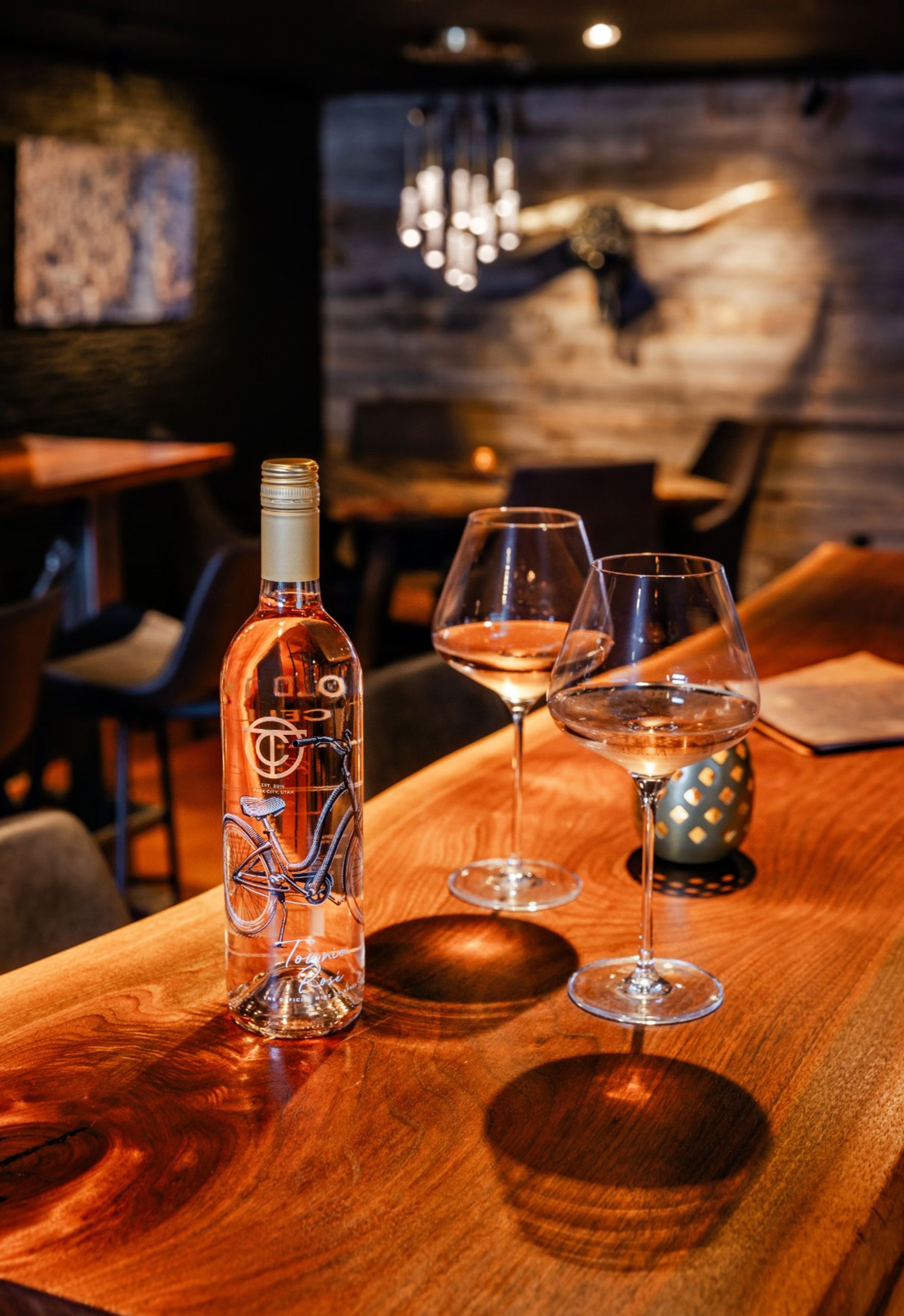 Bottle of rosé with a bicycle label and two poured wine glasses on a polished wooden bar in a cozy, dimly lit wine bar with wood-paneled wall and decorative longhorn in the background.