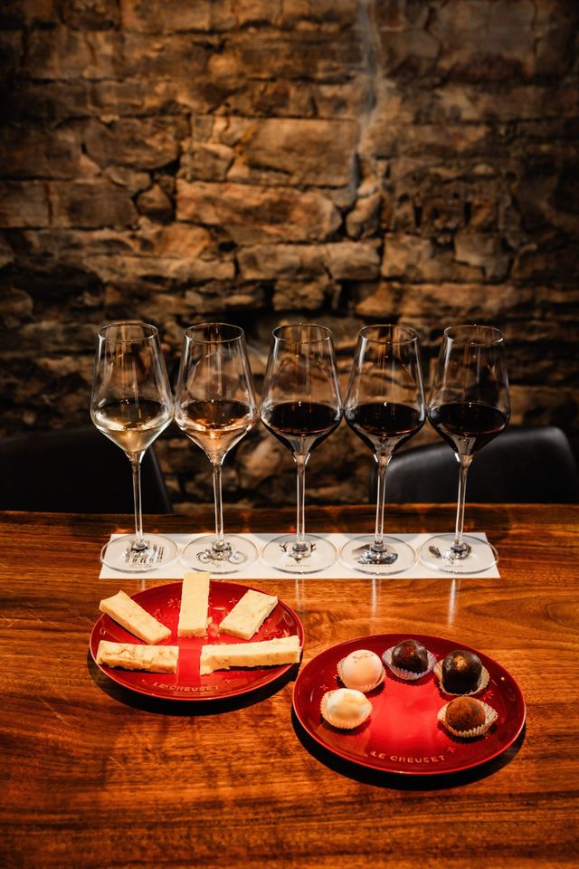 Five-glass wine flight (white, rosé and reds) on a wooden bar against a rustic stone wall, paired with sliced cheese and assorted chocolate truffles on red plates — cozy wine-tasting scene.