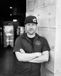Confident warehouse worker in a cap stands with arms crossed, leaning against stacked shipping boxes inside a distribution warehouse (black-and-white photo)
