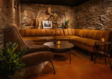 Moody cozy basement lounge with exposed brick and stone walls, caramel leather sectional and suede armchair, round black coffee tables with a wine bottle, two glasses and flickering candles, mounted antler skull and potted plants.