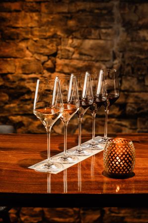 Wine tasting flight of five glasses from rosé to red lined on a wooden bar beside a lit decorative candle against a warm, rustic stone wall in a cozy cellar-style setting.