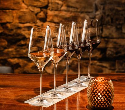 Wine tasting flight of five glasses from rosé to red lined on a wooden bar beside a lit decorative candle against a warm, rustic stone wall in a cozy cellar-style setting.