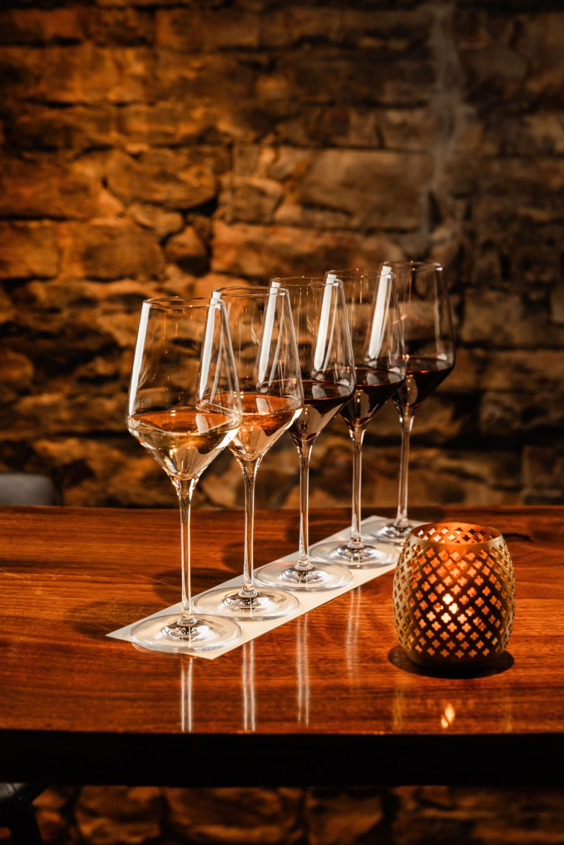 Wine tasting flight of five glasses from rosé to red lined on a wooden bar beside a lit decorative candle against a warm, rustic stone wall in a cozy cellar-style setting.