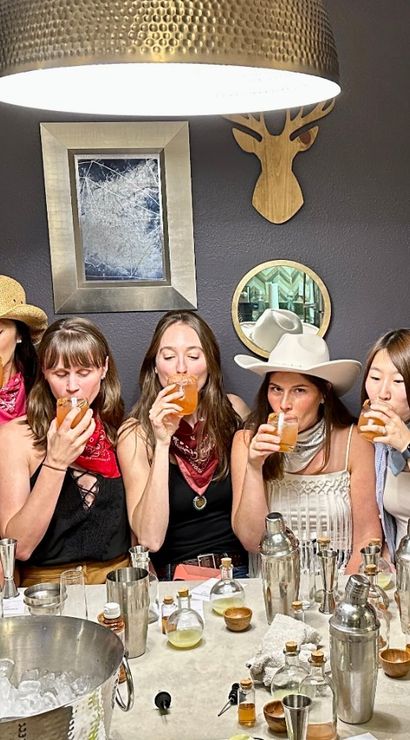 Cowgirl-themed cocktail tasting: a lively group of women sipping drinks around a table with shakers, ice bucket and bar tools in a cozy dining room.