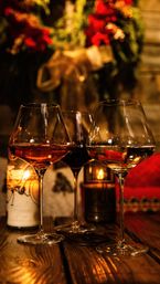 Cozy candlelit trio of wine glasses (red, rosé, white) on a rustic wooden table with a blurred festive wreath and red poinsettias in the background, holiday dining vibe.
