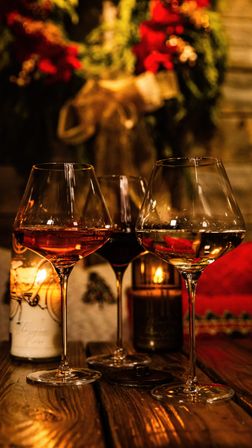 Cozy candlelit trio of wine glasses (red, rosé, white) on a rustic wooden table with a blurred festive wreath and red poinsettias in the background, holiday dining vibe.