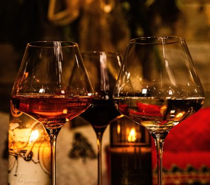 Cozy candlelit trio of wine glasses (red, rosé, white) on a rustic wooden table with a blurred festive wreath and red poinsettias in the background, holiday dining vibe.