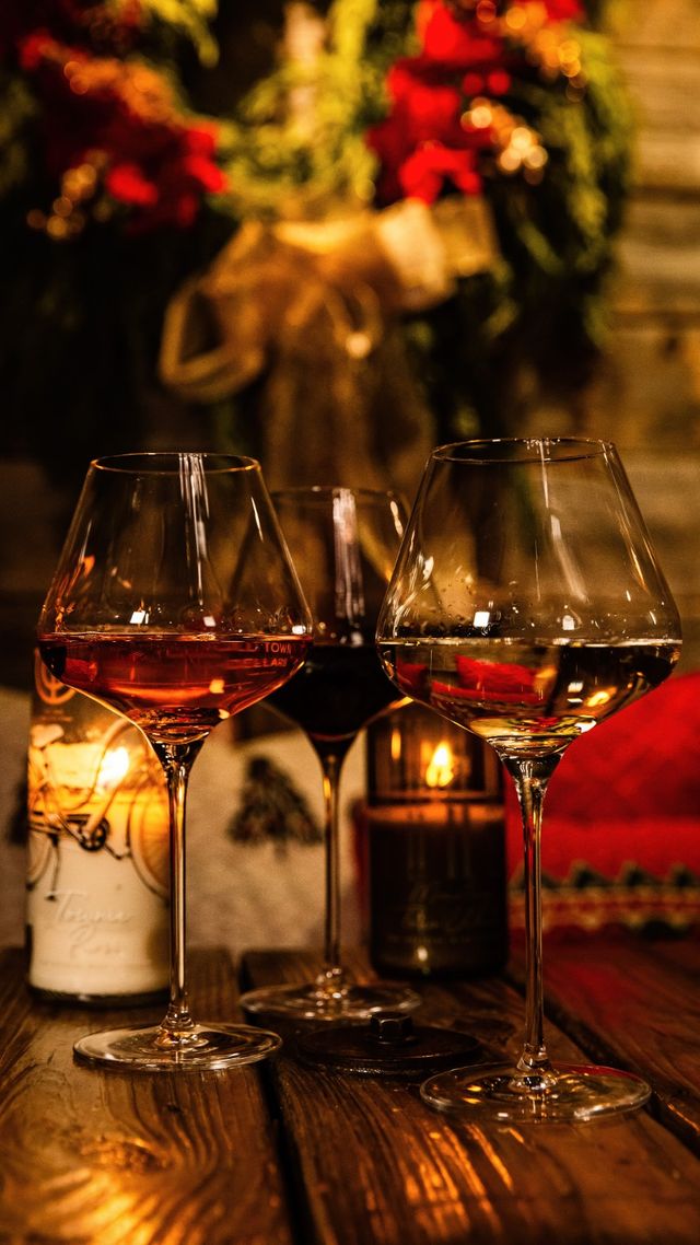 Cozy candlelit trio of wine glasses (red, rosé, white) on a rustic wooden table with a blurred festive wreath and red poinsettias in the background, holiday dining vibe.