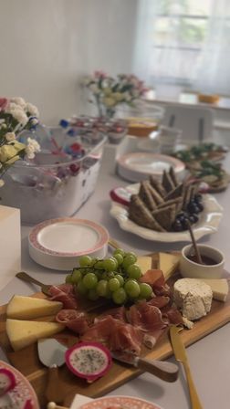 Charcuterie board on a sunlit dining table featuring green grapes, prosciutto, assorted cheeses and goat cheese, a dragon fruit slice, honey pot and crackers with plates and floral centerpieces blurred in the background — elegant brunch spread.
