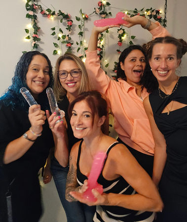 Five women at an indoor bachelorette party smiling and holding pink and clear novelty dildos against a flower-and-fairy-light backdrop.