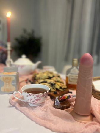 Playful tea table scene with a floral teacup, teapot and pastries, a lit candle and small liquor bottle, plus a pink realistic silicone dildo on a pink cloth.