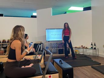 Pilates studio class: instructor stands by a TV presenting while students sit on reformer machines — mats, foam rollers and a snack-and-wine spread in the bright fitness studio.