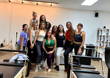 Smiling group of women in activewear posing in a Pilates studio around reformer machines and cubby shelves — upbeat post-class group photo.