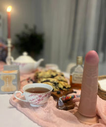 Indoor tea-table still life: floral teacup of tea, pastries on a wooden board, lit taper candle and small bottle, with an upright flesh-toned silicone adult toy on a pink cloth.