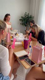 Group of women at a home bachelorette-style party gathered around a table with pink adult novelty favors, notebooks, gift bags, phones and drinks