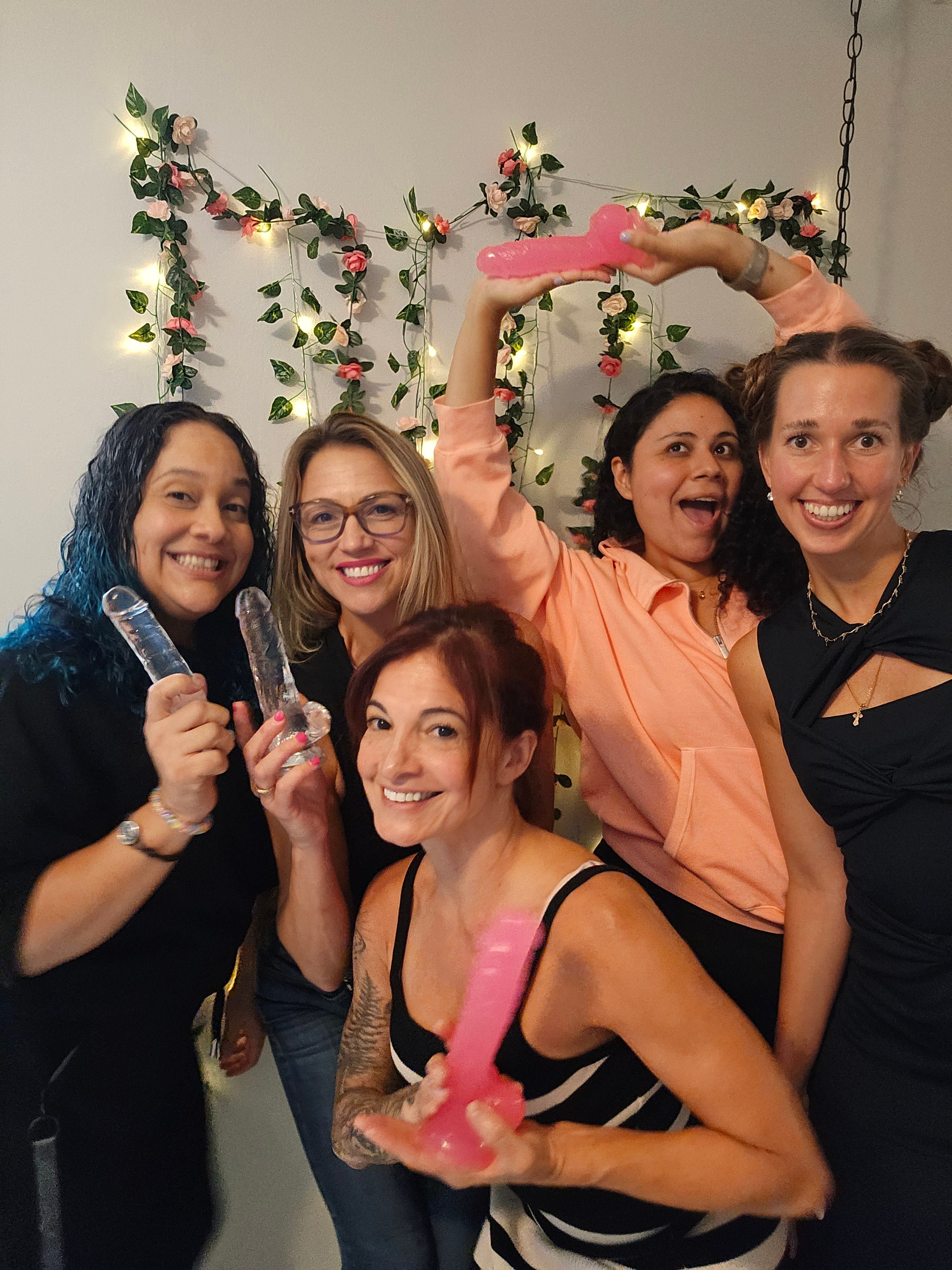Five women at an indoor bachelorette/girls' night smiling and posing while holding colorful silicone dildos in front of a floral fairy-light backdrop