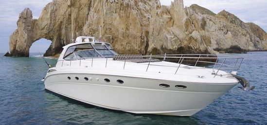 Sleek white luxury motor yacht on turquoise coastal waters beside a dramatic rocky sea arch and rugged cliffs under a cloudy sky