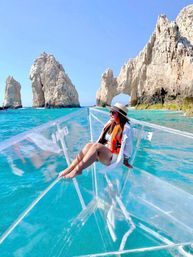 Woman in a sunhat and life jacket sitting on a clear glass-bottom boat over turquoise water, enjoying a sunny view of dramatic limestone rock arches and sea stacks in Los Cabos.