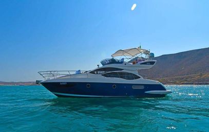 Sleek blue-and-white luxury motor yacht anchored in turquoise coastal waters near sunlit rocky cliffs under a clear blue sky