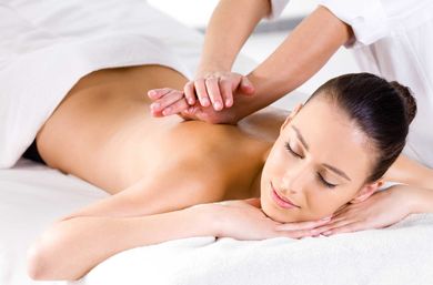 Client lying face down on a white spa table, eyes closed as a therapist's hands deliver a relaxing back massage.