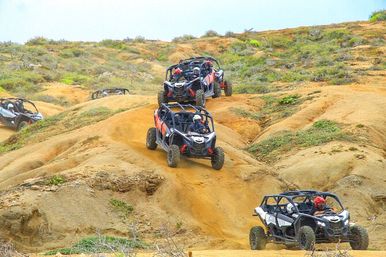 Four off-road UTVs with helmeted drivers tearing down sandy, scrub-covered dunes on a rugged desert trail — adrenaline-filled dune buggy adventure
