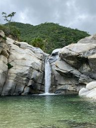 Small waterfall cascading between smooth granite boulders into a clear green swimming pool, set in a rocky gorge with lush hillside vegetation under a cloudy sky.