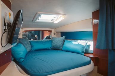 Cozy yacht cabin v-berth with teal bedding and pillows, wood trim, wall-mounted TV, striped accent pillow and overhead hatch skylight.