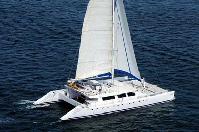 Aerial view of a white luxury catamaran with twin hulls and a tall white sail, cruising on deep blue open ocean with a blue sail cover.