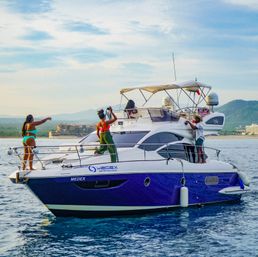 Friends enjoying a sunny day on a purple-and-white motor yacht in calm blue coastal waters with a distant shoreline and green hills, taking photos and relaxing on deck