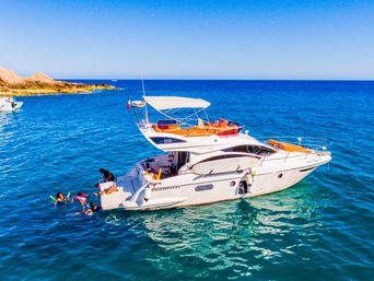 Sunny white luxury motor yacht anchored in clear turquoise coastal waters near a rocky shoreline, people in colorful snorkels and life vests swimming and playing beside the boat under a bright blue sky