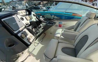 Sunlit motorboat helm and cockpit with white leather captain seats, steering wheel and instrument dashboard, docked at a busy marina with colorful boats in the background.