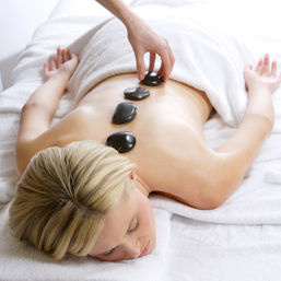 Relaxing hot stone massage at a spa — smooth black basalt stones lined along a person’s bare back as a therapist places one, surrounded by white towels and linens.