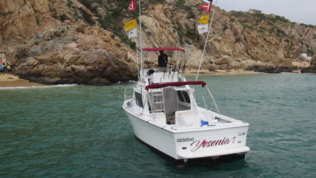 White sportfishing boat with red canopy and fishing rods in turquoise water beside a rocky coastline and small sandy beach