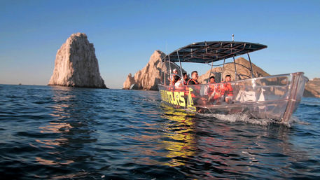 Clear-hulled tour boat with passengers in orange life jackets cruising past dramatic coastal rock formations at golden hour on a calm blue ocean.