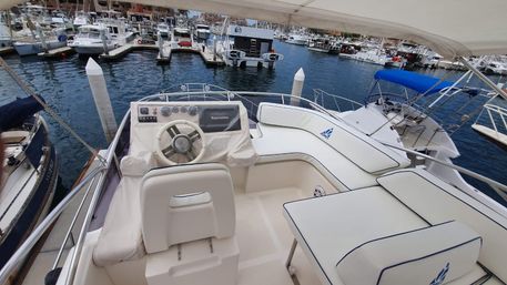 White cushioned yacht flybridge with helm, steering wheel and navigation display, overlooking a busy marina with docked boats and calm harbor waters.