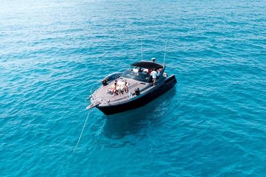 Aerial view of a sleek black motorboat anchored in clear turquoise ocean, people sunbathing on the bow and relaxing under the shaded cockpit.