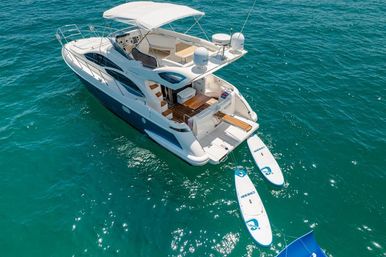 Aerial view of a white luxury motor yacht on clear turquoise water with a shaded flybridge, open teak swim platform and two tethered paddleboards ready for fun