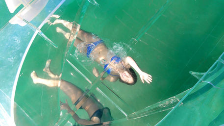 Person in a blue bikini floating inside a clear inflatable water sphere on turquoise open water, overhead view with mirrored reflections